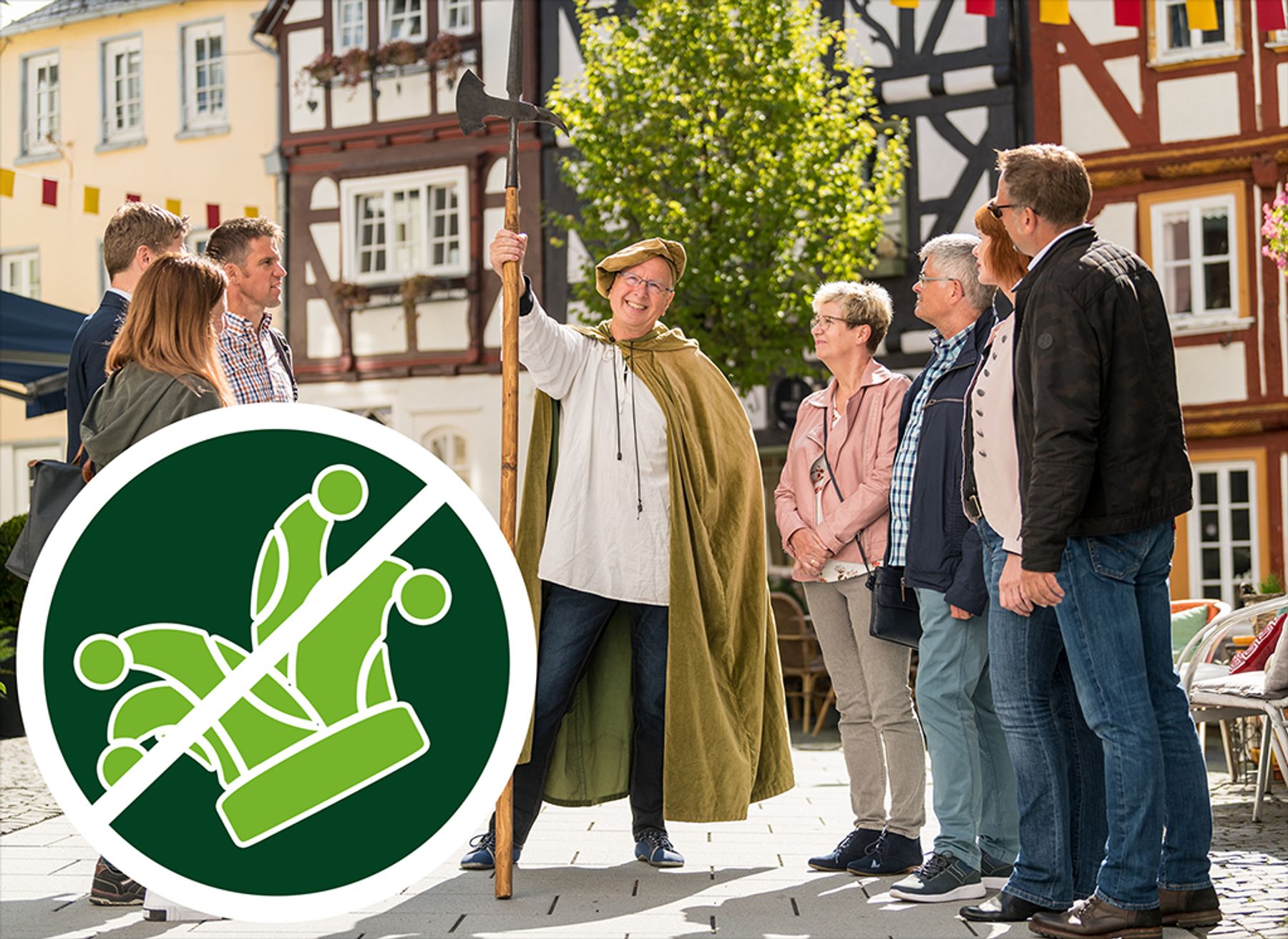 Eine Person in mittelalterlicher Tracht hält einen Stab und lächelt, während sie zu einer Gruppe leger gekleideter Erwachsener im Freien spricht, hinter denen sich Fachwerkhäuser befinden. Im Vordergrund ist eine "Keine Krone"-Grafik eingeblendet.